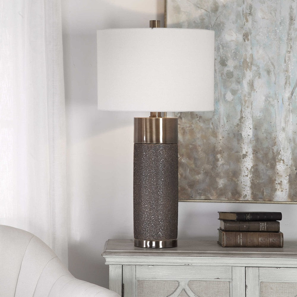 This contemporary table lamp design showcases a heavily textured ceramic base that is finished in a metallic golden bronze, paired with antique brass plated iron details. A hardback drum shade in an off-white linen fabric completes this piece.