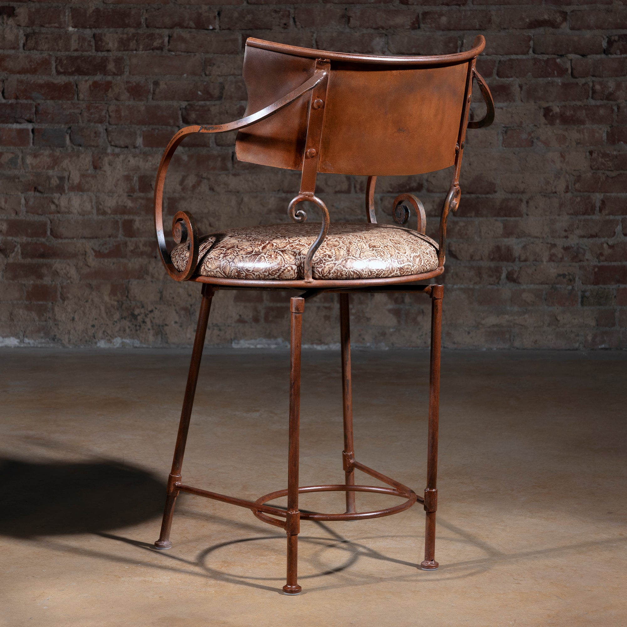 Back view of copper-toned metal bar stool with forged iron frame and curved scroll arms