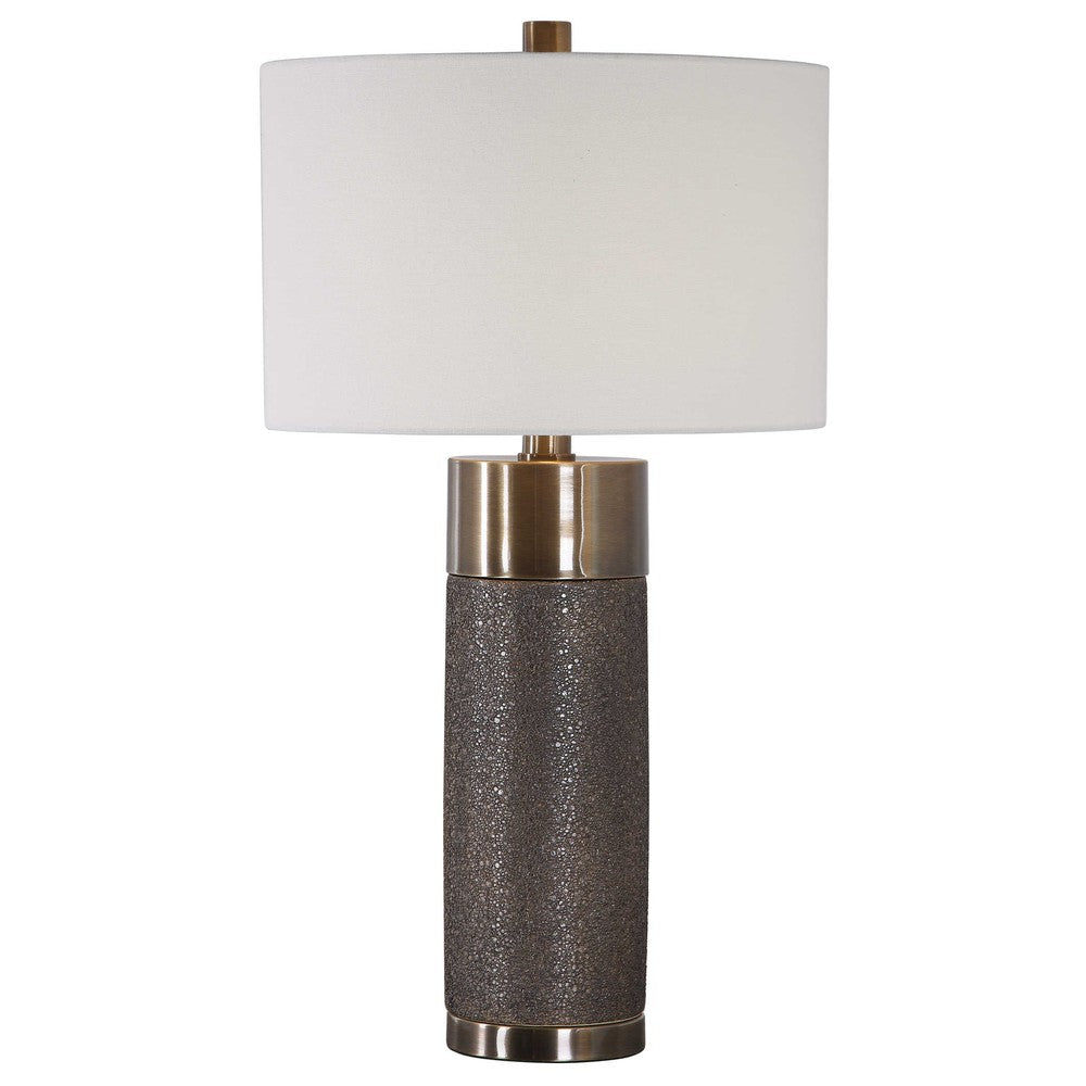 This contemporary table lamp design showcases a heavily textured ceramic base that is finished in a metallic golden bronze, paired with antique brass plated iron details. A hardback drum shade in an off-white linen fabric completes this piece.