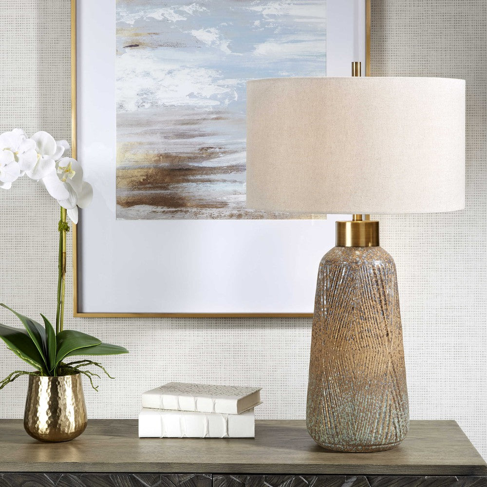 A beautiful display of color and style, this ceramic table lamp features an embossed design with a unique mix of matte and gloss glazes in various shades of blue-green, indigo, and golden rust, paired with antique brass plated details. The hardback drum shade is covered in a light gray linen fabric with natural slubbing.