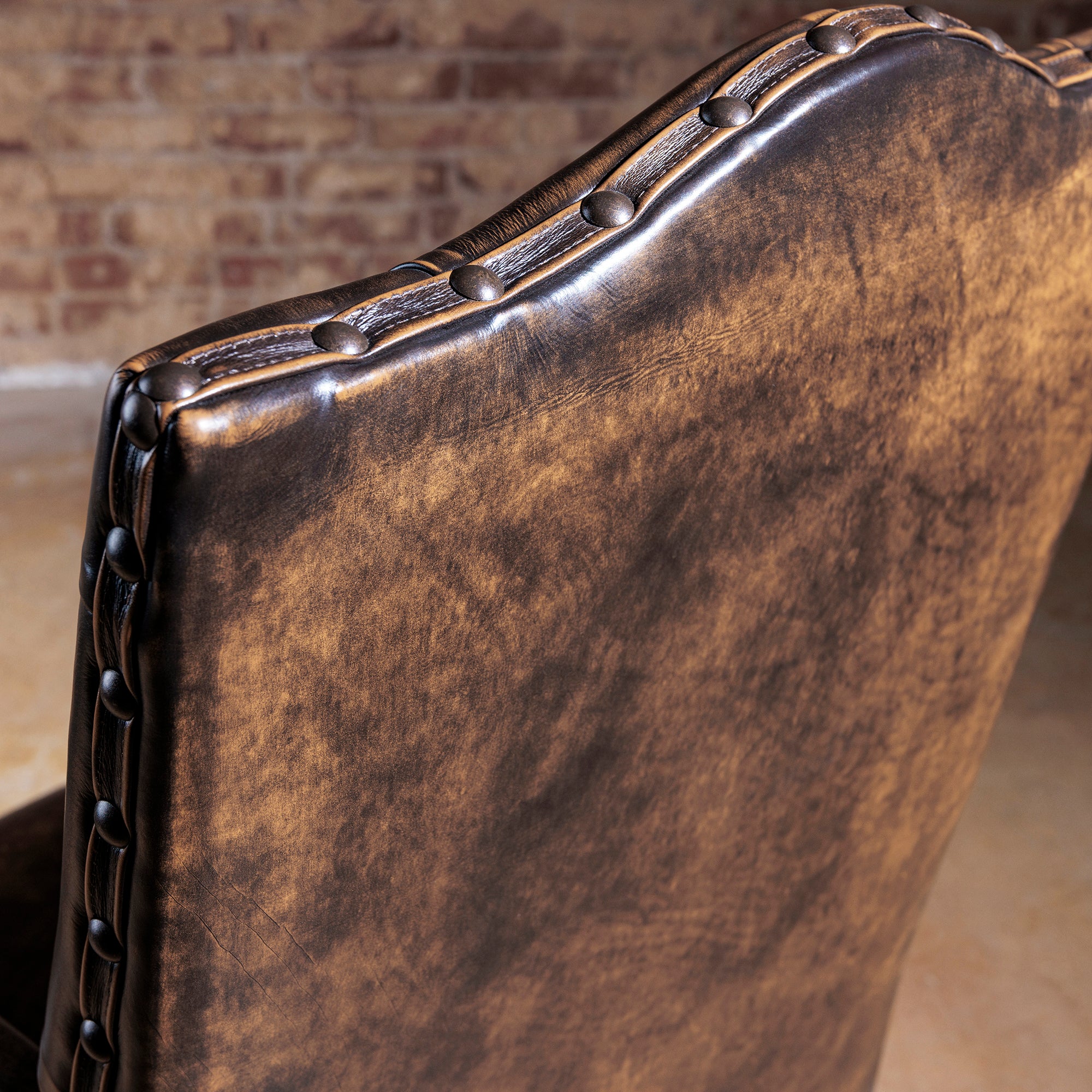 Close-up of nailhead trim along the top edge of a distressed brown leather high-back Western arm chair.