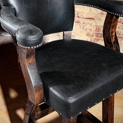Close-up of black leather seat and armrest with hand-applied nailhead trim on wood frame.