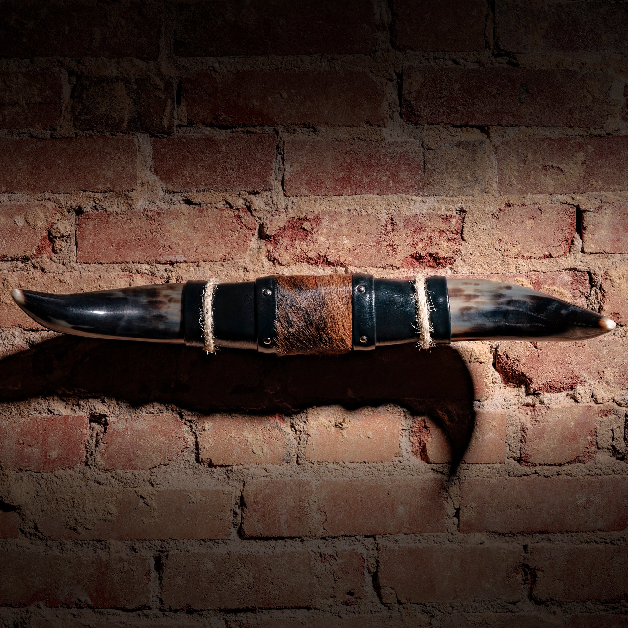 Mounted longhorn horn decor with cowhide center, black leather ends, and jute rope accents on brick wall