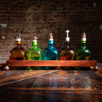 Set of five colored glass bottles with decorative tops in a copper tray on a rustic wood table