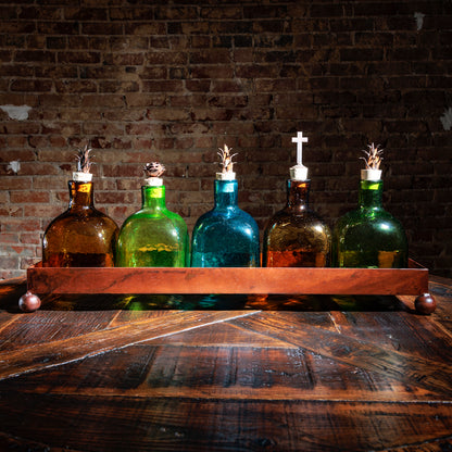Set of five colored glass bottles with decorative tops in a copper tray on a rustic wood table