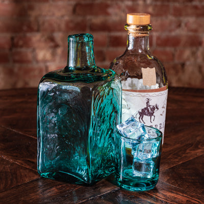 Shot glass filled with ice next to matching teal whiskey decanter and liquor bottle